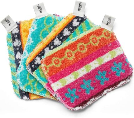 Four assorted color Skoy Scrubs, round with various patterns, arranged on a clean kitchen counter next to a sink.