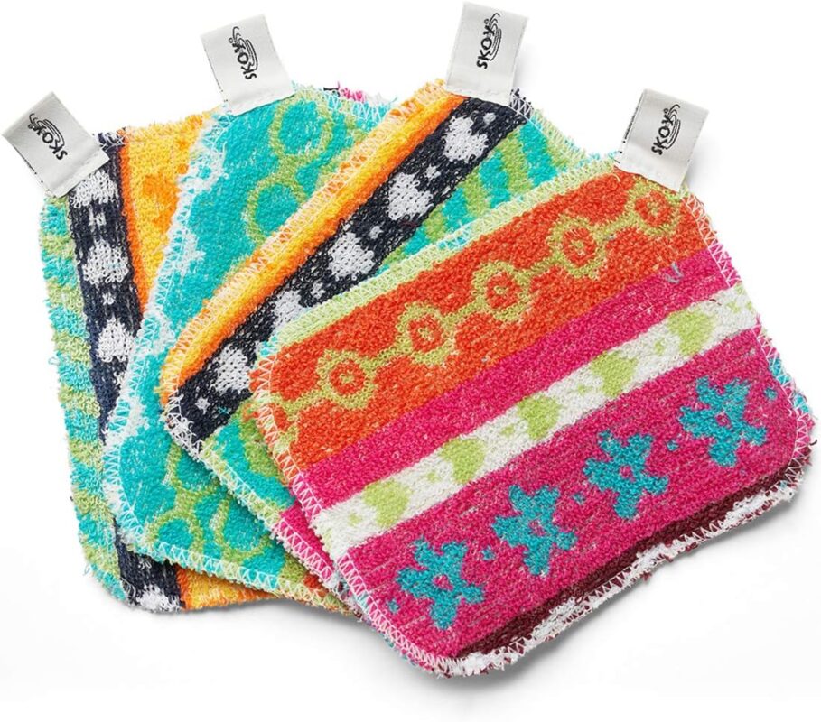 Four assorted color Skoy Scrubs, round with various patterns, arranged on a clean kitchen counter next to a sink.